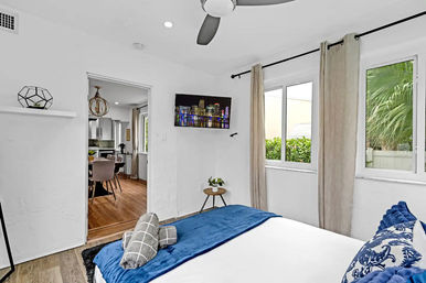 Bright, cozy bedroom in a vacation rental with blue-accent bedding, ceiling fan, wall-mounted TV showing a city skyline, beige curtains over windows looking onto tropical greenery, and a doorway to a dining area with wood floors.