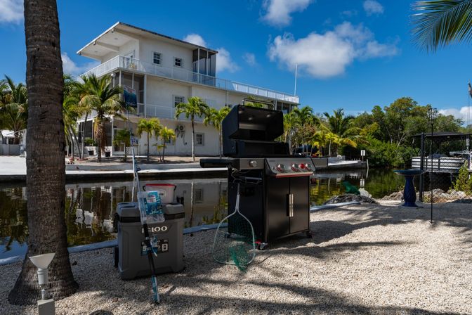 Tropical canal-side scene: black gas grill, cooler and fishing net on a gravel patio with palm trees and a white waterfront building under a bright blue sky.