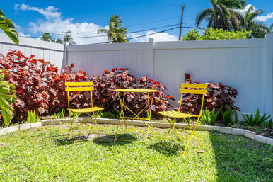 Sunny tropical backyard with a bright yellow bistro table and two chairs on a green lawn, red-leaf shrubs and palm trees behind a white privacy fence.