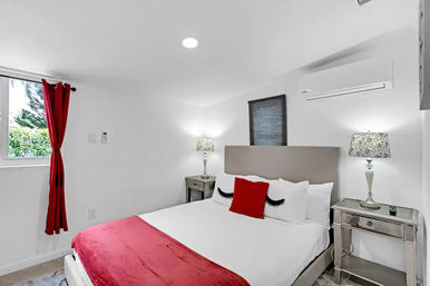 Bright modern bedroom with gray upholstered headboard, white bedding, red throw and red accent pillow, decorative eyelash pillows, mirrored nightstands with patterned lamps, red curtains at a window showing greenery, and a wall-mounted air conditioner.