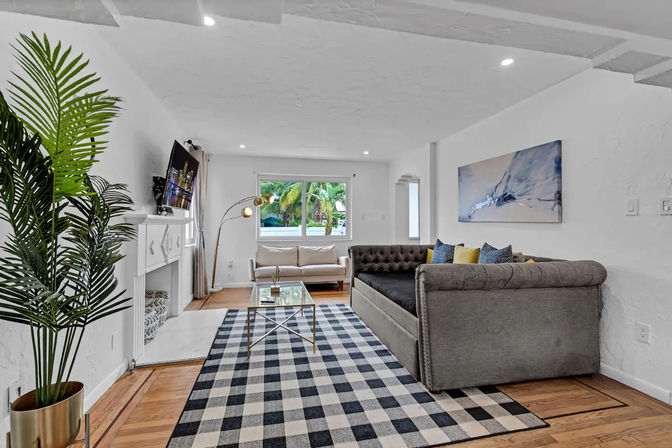 Sunlit modern tropical living room with tufted gray sofa, beige loveseat, black-and-white checkered rug, glass coffee table, wall-mounted TV, potted palm and window view of palm trees.