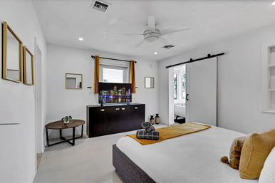 Bright modern vacation-rental bedroom with white walls, ceiling fan, sliding barn door, mustard-yellow accents, TV showing a city skyline, round wood side table, and neatly made bed with rolled towels.