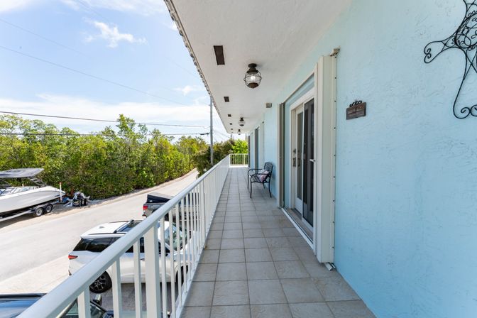 Sunny second-floor balcony of a light-blue coastal condo with tiled walkway and white railing, wall lanterns, a metal bench by sliding glass doors, parked cars and a trailer boat on the street, green trees and blue sky beyond.