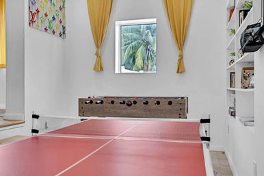 Home game room with red ping-pong table in the foreground, wooden foosball table against a white wall, small square window framing tropical palm trees, tied-back yellow curtains, built-in white shelves and colorful wall art.