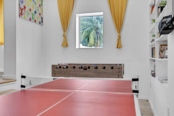 Home game room with red ping-pong table in the foreground, wooden foosball table against a white wall, small square window framing tropical palm trees, tied-back yellow curtains, built-in white shelves and colorful wall art.