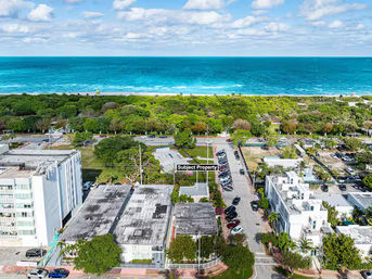 Aerial view of a near-beach coastal city block with low-rise buildings, parked cars and a labeled 'Subject Property' lot, tree-lined street and green park strip leading to a turquoise ocean and sandy beach on the horizon.