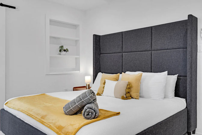 Bright modern bedroom with tall gray upholstered headboard, white linens, mustard-yellow throw and pillows, rolled gray towels on the bed, built-in white shelves and a bedside lamp — cozy minimalist guest room.