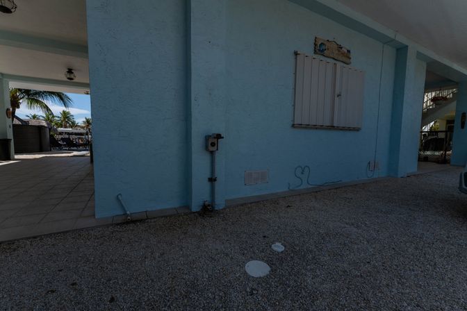 Light-blue stucco coastal house with covered carport and gravel driveway, closed window shutter and utility box, tiled patio opening to palm trees and pool loungers in the background.