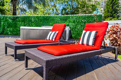 Two wicker sun loungers with bright red cushions and black-and-white striped pillows on a sunny wooden patio deck, green hedge and tropical plants in the background.