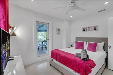 Bright modern white bedroom with king bed dressed in fuchsia throw and pillows, rolled gray towels, ceiling fan, wall art and TV, with glass door opening to a covered balcony and tropical palm view.
