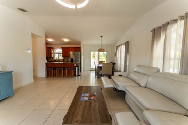 Sunlit open-concept living room and kitchen with beige tile floors, cream leather reclining sofa, wooden coffee table with magazine, kitchen island and bar stools, dining area by sliding glass doors revealing palm trees outside.