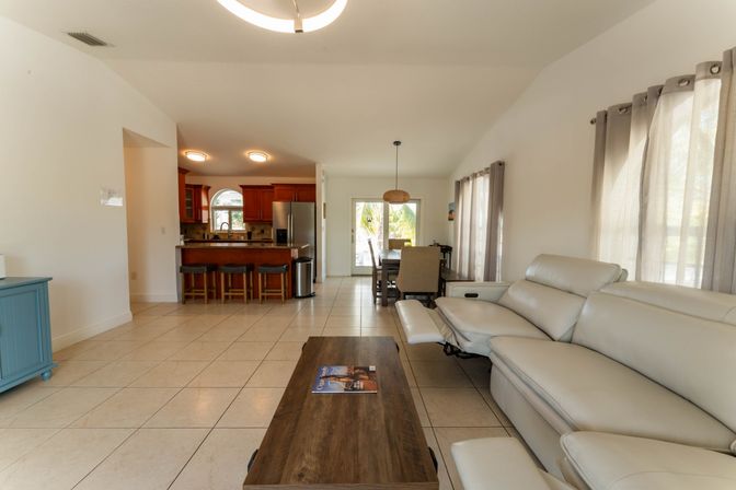 Sunlit open-concept living room and kitchen with beige tile floors, cream leather reclining sofa, wooden coffee table with magazine, kitchen island and bar stools, dining area by sliding glass doors revealing palm trees outside.