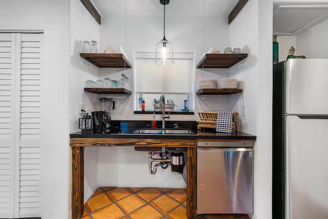 Cozy kitchen sink nook with black countertop, farmhouse-style wooden open shelves displaying dishes and glasses, stainless-steel dishwasher, pendant light, coffee maker, and terracotta tile floor.