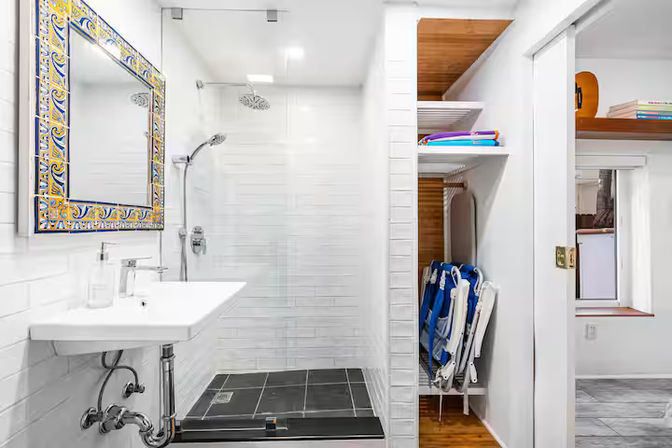 Bright modern white bathroom with walk-in glass shower, black tiled shower floor, decorative blue-and-yellow framed mirror, wall-mounted sink, and open shelving holding folded towels and beach-ready blue folding chairs.