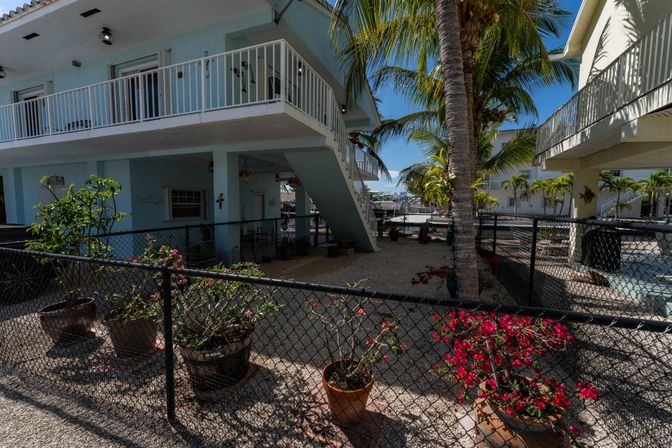 Tropical waterfront two-story condo with white balcony and exterior stairs, palm tree and potted pink bougainvillea in a gravel courtyard behind a black chain-link fence, marina boats visible in the background