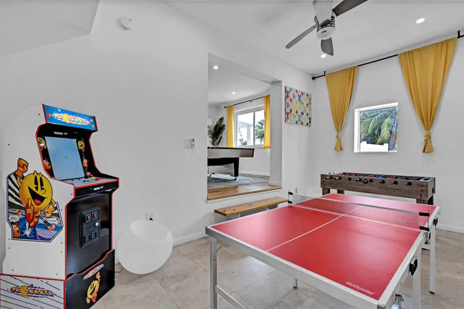 Bright game room with red ping-pong table, retro arcade cabinet featuring Pac-Man artwork, foosball table, yellow curtains, ceiling fan and palm trees visible through the window