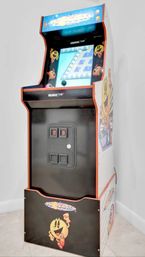 Retro Pac-Man arcade cabinet — upright arcade machine with colorful marquee, joystick and game screen, black-and-white cabinet featuring smiling Pac-Man artwork standing against a white wall on a tiled floor