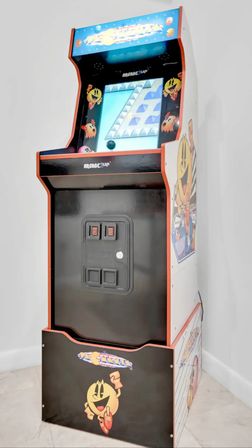 Retro Pac-Man arcade cabinet — upright arcade machine with colorful marquee, joystick and game screen, black-and-white cabinet featuring smiling Pac-Man artwork standing against a white wall on a tiled floor