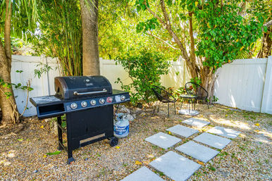 Sunny tropical backyard patio with a black gas grill and propane tank on gravel, concrete stepping stones leading to a small seating area with two wire chairs and table, surrounded by lush trees and a white privacy fence.
