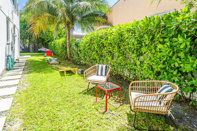 Sunny tropical backyard with a palm tree and dense green hedge, two wicker lounge chairs with striped cushions and a small red side table on a grassy lawn, stepping stones and lawn games in the distance.
