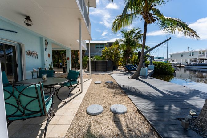 Waterfront tropical patio with teal-cushioned dining set, palm trees, wooden dock and boats along a calm canal under a bright blue sky.