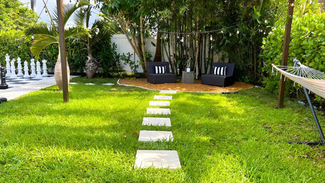 Tropical backyard lawn with square stepping stones leading to a cozy seating nook of black outdoor chairs under string lights, surrounded by palm and bamboo, hammock and oversized outdoor chess pieces.