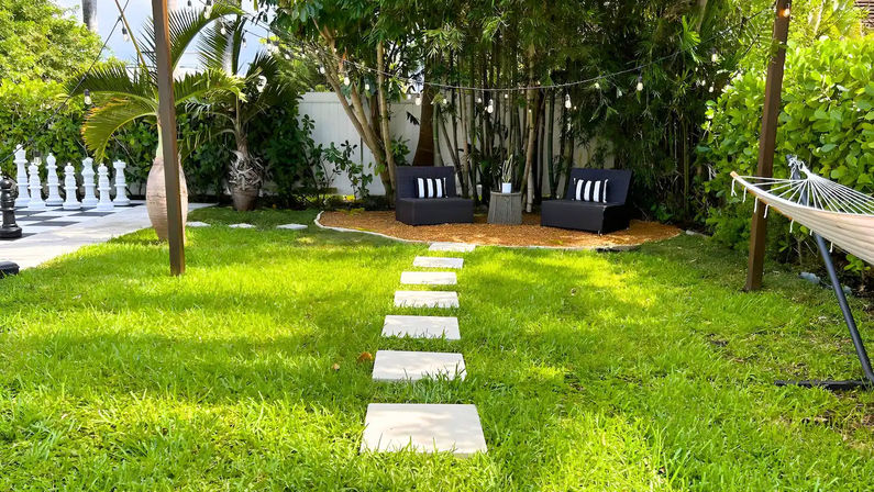 Tropical backyard lawn with square stepping stones leading to a cozy seating nook of black outdoor chairs under string lights, surrounded by palm and bamboo, hammock and oversized outdoor chess pieces.