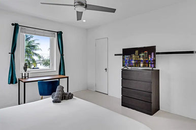 Sunlit modern minimalist coastal bedroom — teal curtains framing a palm-tree window, compact desk with blue chair, ceiling fan, dark dresser with TV showing a city skyline, rolled towels on the bed.