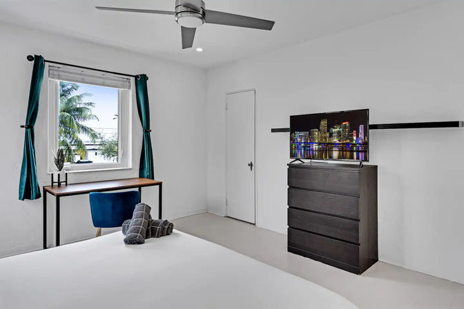 Sunlit modern minimalist coastal bedroom — teal curtains framing a palm-tree window, compact desk with blue chair, ceiling fan, dark dresser with TV showing a city skyline, rolled towels on the bed.