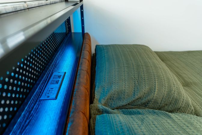 Modern bedroom detail — dark wood and leather headboard with built-in power outlets and blue LED lighting beside teal textured pillows and quilt.