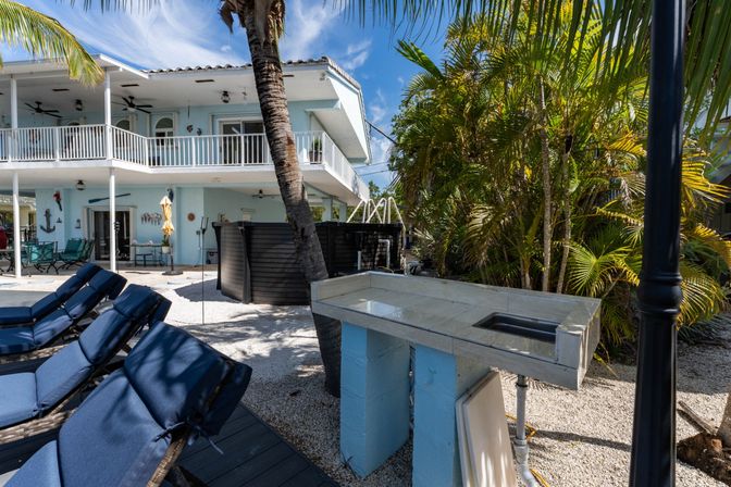 Sunlit coastal backyard with a light-blue two-story house and balcony, navy cushioned lounge chairs, palm trees, above-ground pool, tiled outdoor sink and white gravel patio — tropical Florida-style setting