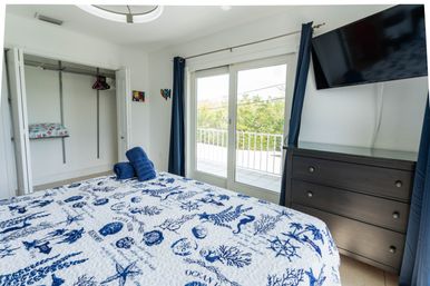 Coastal-themed bedroom with blue-and-white ocean-print quilt, rolled navy towels on the bed, open closet, sliding glass balcony doors showing greenery, dark wood dresser and wall-mounted TV.
