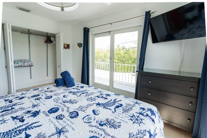 Coastal-themed bedroom with blue-and-white ocean-print quilt, rolled navy towels on the bed, open closet, sliding glass balcony doors showing greenery, dark wood dresser and wall-mounted TV.