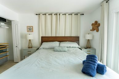 Cozy coastal bedroom retreat with a king bed, woven wood headboard, pale blue linens, rolled navy towels at the foot, matching nightstands and lamps, neutral curtains and beach-themed wall decor.