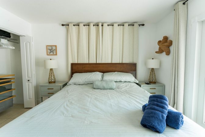 Cozy coastal bedroom retreat with a king bed, woven wood headboard, pale blue linens, rolled navy towels at the foot, matching nightstands and lamps, neutral curtains and beach-themed wall decor.