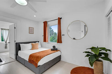 Sunlit coastal bedroom with gray upholstered bed, white linens, rust-orange throw and curtains, round gold mirror, potted plant, and window view of palm trees.