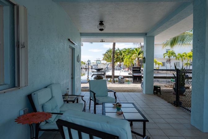Covered canal-front patio with light-blue cushioned seating and coffee table, overlooking a docked boat, palm trees and a tropical waterfront with a barbecue grill.