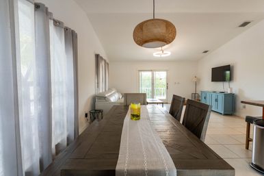Light-filled open-plan dining and living room with a rustic wood dining table and beige runner, yellow glass centerpiece, white sofa, sliding glass doors, blue media console and rattan pendant light.