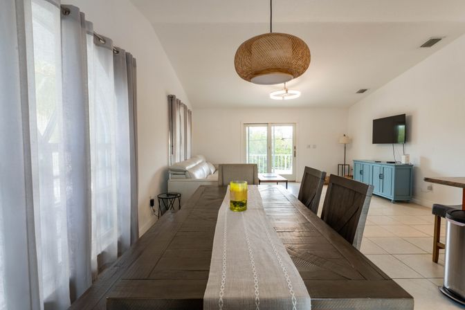 Light-filled open-plan dining and living room with a rustic wood dining table and beige runner, yellow glass centerpiece, white sofa, sliding glass doors, blue media console and rattan pendant light.
