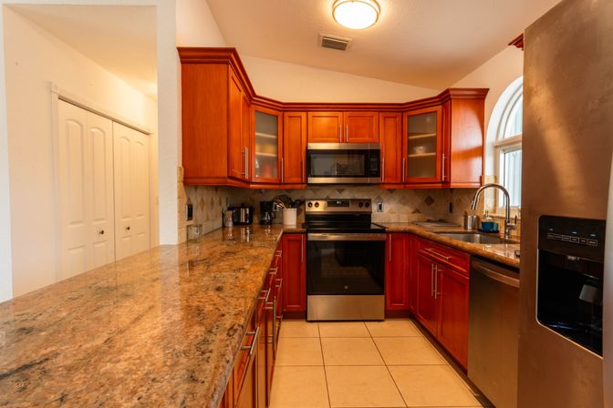 Warm U-shaped residential kitchen with cherry wood cabinets, speckled granite countertops, stainless steel oven, microwave and refrigerator, tile backsplash and floor, and a sink under an arched window.