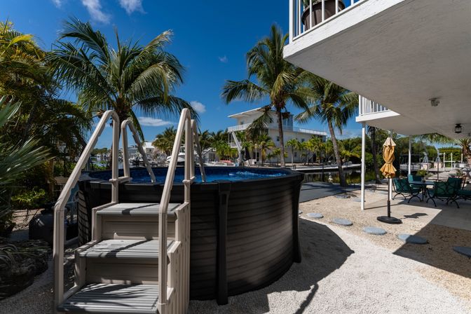 Sunny tropical waterfront backyard with round above-ground pool and ladder, palm trees along a canal, and shaded patio dining under a raised white balcony