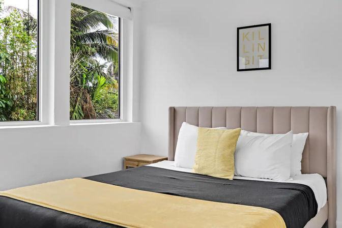 Bright minimalist bedroom with a beige upholstered headboard, black-and-yellow bedding and large windows revealing lush tropical palm trees outside.