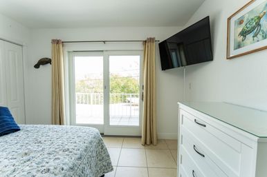 Sunny coastal bedroom with patterned quilt and blue towels, sliding glass doors to a balcony, wall-mounted TV, white dresser and sea turtle artwork.