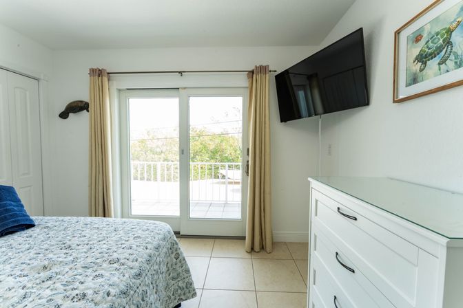 Sunny coastal bedroom with patterned quilt and blue towels, sliding glass doors to a balcony, wall-mounted TV, white dresser and sea turtle artwork.
