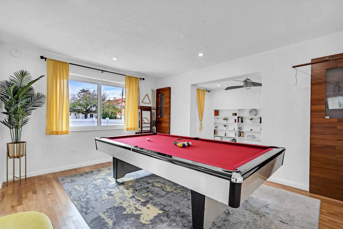 Bright modern game room with red-felt pool table, hardwood floors, large window with sunny yellow curtains, built-in white bookshelves, ceiling fan and potted palm in a stylish residential interior