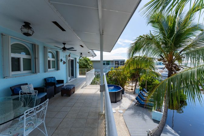 Tropical waterfront balcony with light-blue exterior, outdoor wicker seating and glass dining table, ceiling fans, palm trees, dockside lounge chairs, small round hot tub and marina view