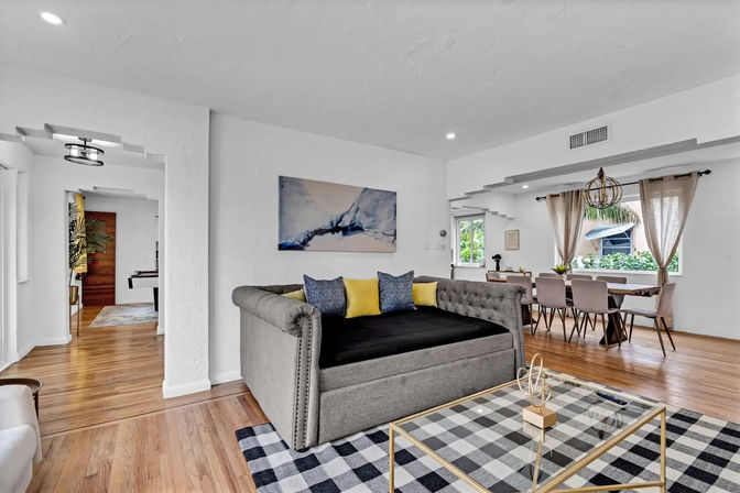 Sunlit open-concept living and dining room interior with hardwood floors, tufted gray daybed sofa topped with yellow and patterned pillows, black-and-white checkered rug, glass coffee table, and a dining table by large windows with beige curtains and a decorative pendant light.