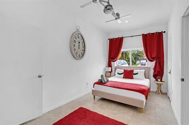 Bright modern bedroom with pops of red: bed with eyelash pillow and red throw, matching curtains, wall clock, ceiling fan, tiled floor, and window view of palm trees and parked cars.