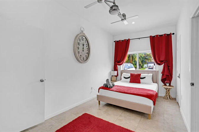Bright modern bedroom with pops of red: bed with eyelash pillow and red throw, matching curtains, wall clock, ceiling fan, tiled floor, and window view of palm trees and parked cars.