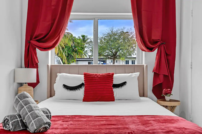 Cozy, chic bedroom with bold red curtains and bed runner, tufted headboard, eyelash-print pillows with a ruffled red cushion, rolled gray towels on the bed and a window framing palm trees and a leafy tree outside.